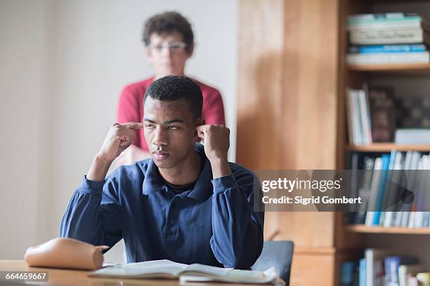 young male high school student sitting at desk with fingers in ears and teacher standing behind - vingers in de oren stockfoto's en -beelden