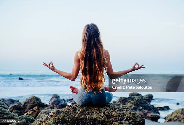 rear view of young woman with long hair practicing lotus yoga pose on rocks at beach, los angeles, california, usa - lotus position stock pictures, royalty-free photos & images