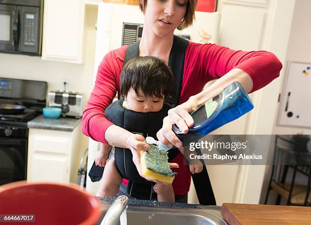 mother with baby in carrier, washing dishes - detergente líquido fotografías e imágenes de stock