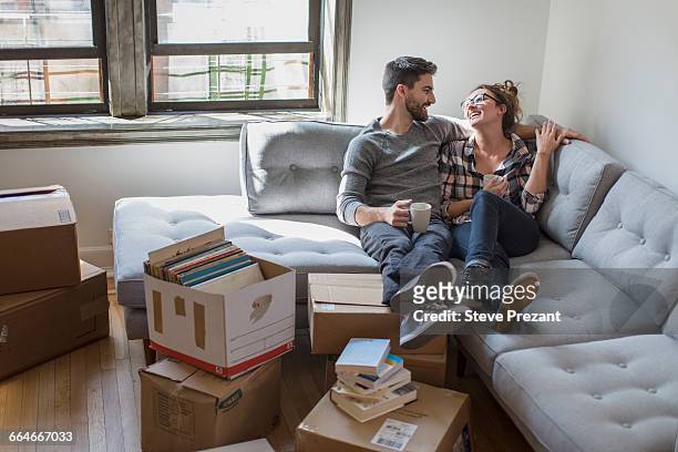 moving house: young couple relaxing on sofa surrounded by cardboard boxes - young adult couple stock pictures, royalty-free photos & images