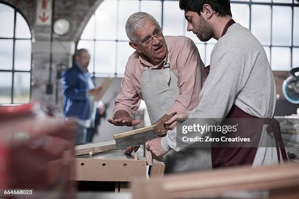 senior carpenter explaining sawing to trainee in workshop - werk in uitvoering stockfoto's en -beelden