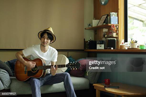 young man playing guitar on sofa in living room - man fedora room stock pictures, royalty-free photos & images