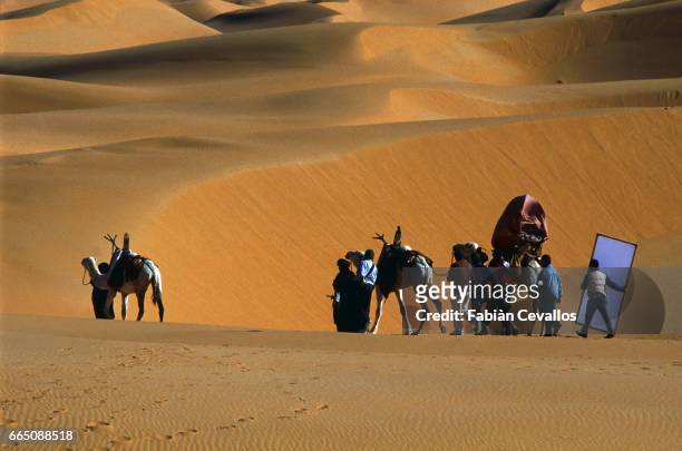 Bernardo Bertolucci and his crew carry equipment and follow a camel rider, on the set of the movie Un The au Sahara, or Il Te Nel Deserto, originally...