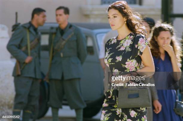 Italian actress Monica Bellucci, wearing a black dress with flowers and carrying a black purse, walks down the streets with two men with guns in the...