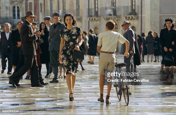 Italian actress Monica Bellucci, wearing a black dress with flowers and carrying a black purse, crosses a wet square with passersby, a police officer...
