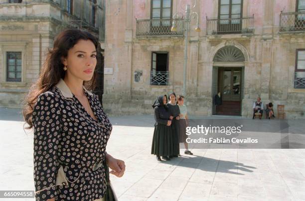 Italian actress Monica Bellucci, wearing a black dress, stands on a square with three women walking behind her and a classical building in the...