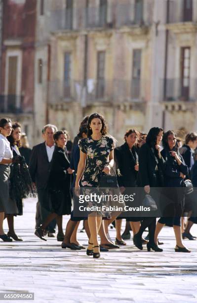 Italian actress Monica Bellucci, wearing a black dress with flowers and carrying a black purse, crosses a wet square with a group of mourning women...