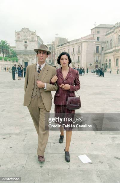 Italian actress Monica Bellucci walks across a square, holding arms with a man wearing a suit and a hat, with classical buildings in the background...