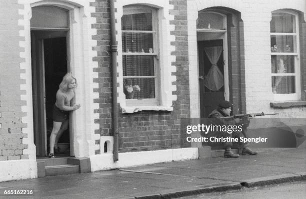 Woman leans out of her doorway as a soldier aims his rifle down a Belfast street, 1972. Belfast residents suffer from continuous street battles in...