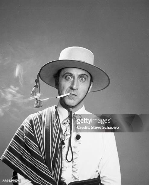 Actor Gene Wilder dressed in costume as the silent screen star Rudolph Valentino, 1977. He is smoking a cigarette and wearing a hat, mimicking...