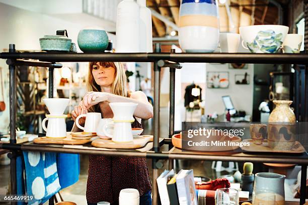 woman standing in a shop, arranging ceramic objects on a shelf. - tienda de regalos fotografías e imágenes de stock