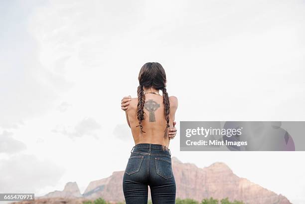 rear view of a topless young woman with long brown hair in braids, wearing jeans, tattoo of a celtic cross on her back. - cruz celta imagens e fotografias de stock