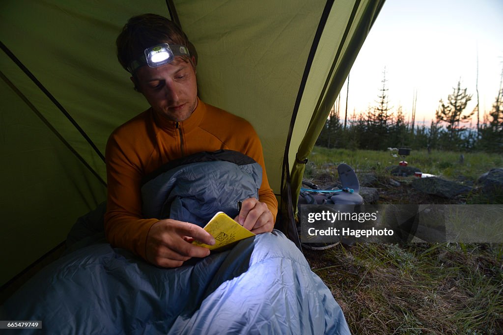 Male camper in sleeping bag writing his journal at dusk on Midnight Ridge, Colville National Forest, Washington State, USA