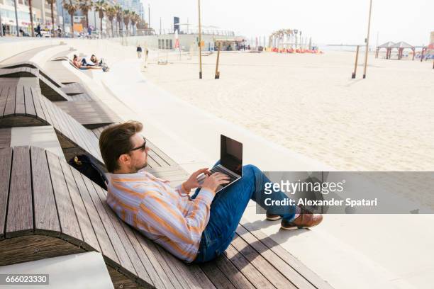 man using laptop sitting on a wooden sunbed on the beach - israel travel stock pictures, royalty-free photos & images