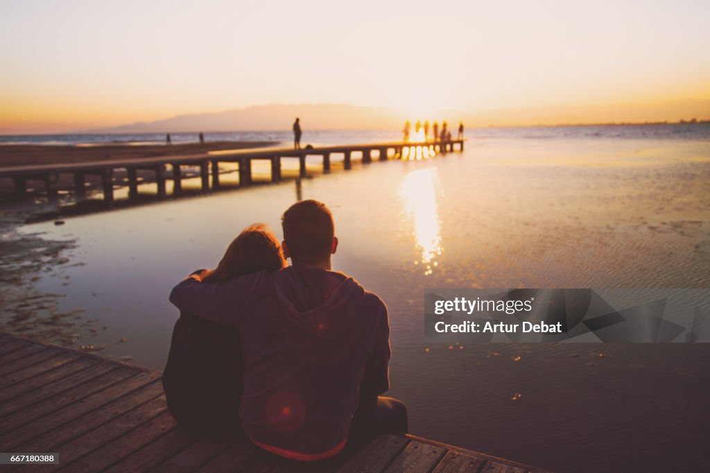 Couple in love during romantic weekend traveling together, sitting in the wood pier contemplating the sunset over the Ebro Delta in the Mediterranean Sea with beautiful colors.