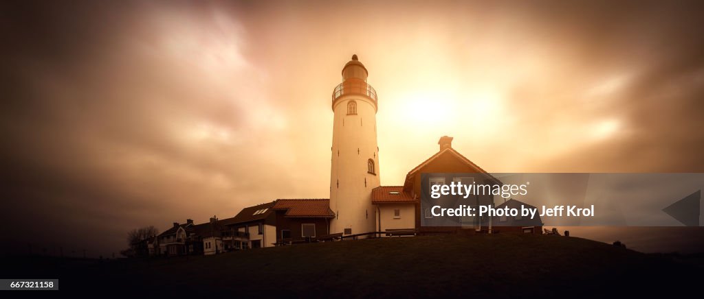 Panoramic view of a lighthouse on a hill with houses, windy sky with sun coming through the clouds.