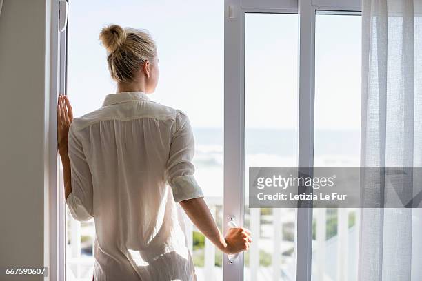 young woman standing by window at home - bay window interior stock pictures, royalty-free photos & images