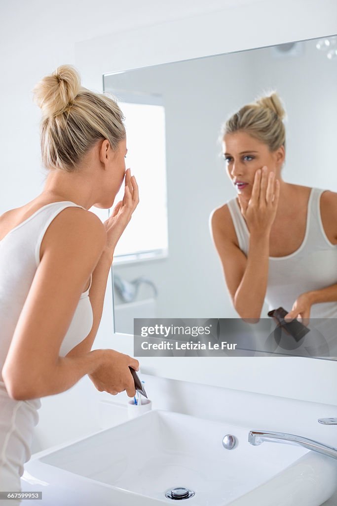 Beautiful young woman applying moisturizer on her face in bathroom
