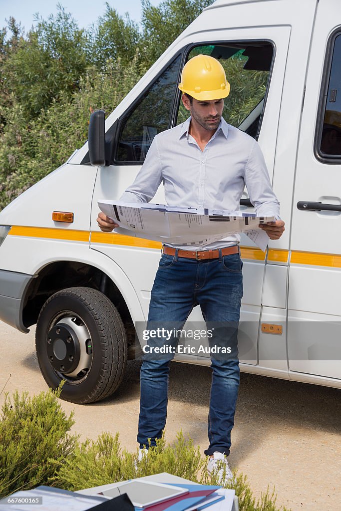 Male engineer reading a blueprint by van at site