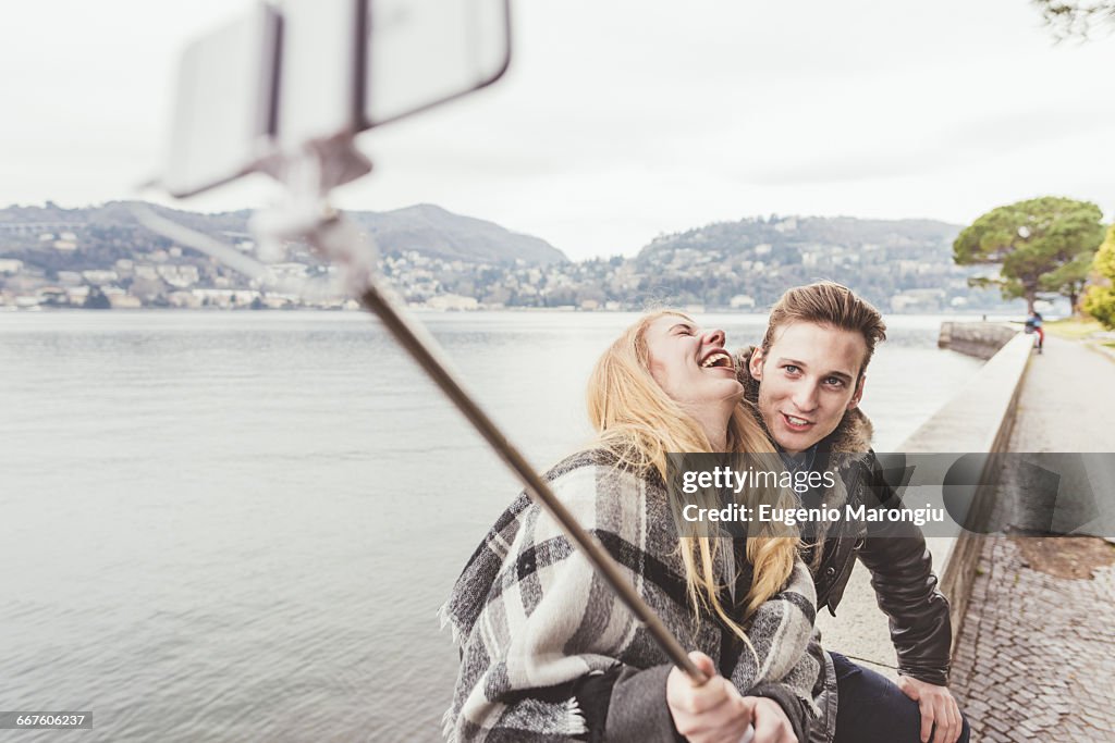 Young couple laughing taking smartphone selfie on harbour wall, Lake Como, Italy