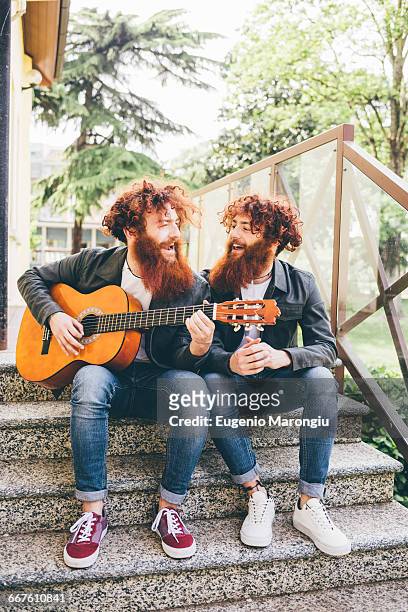 young male hipster twins with red beards sitting on stairway playing guitar - man singing stockfoto's en -beelden