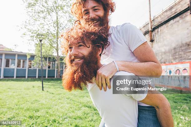 young male hipster twins with red beards piggy backing in park - twin fotografías e imágenes de stock