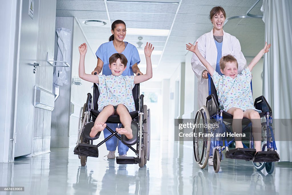 Medical orderlies having fun pushing boy patients in wheelchair on hospital childrens ward