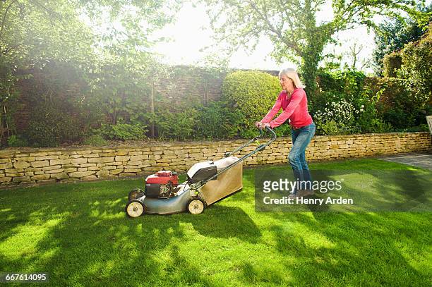 mature woman mowing sunlit garden lawn with lawn mower - mowing lawn stock-fotos und bilder
