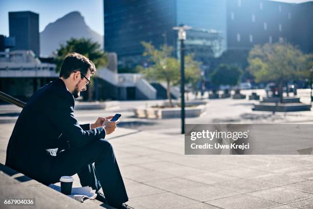 young businessman texting while sitting on stairs - vista traseira a três quartos - fotografias e filmes do acervo