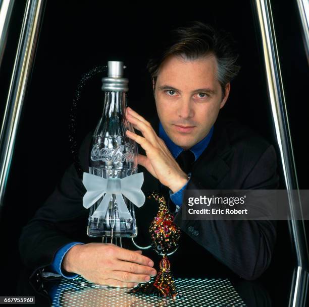 French fashion designer Olivier Lapidus holds his crystal perfume atomizer shaped like a Coca-Cola bottle. French embroiderer Francois Lesage created...