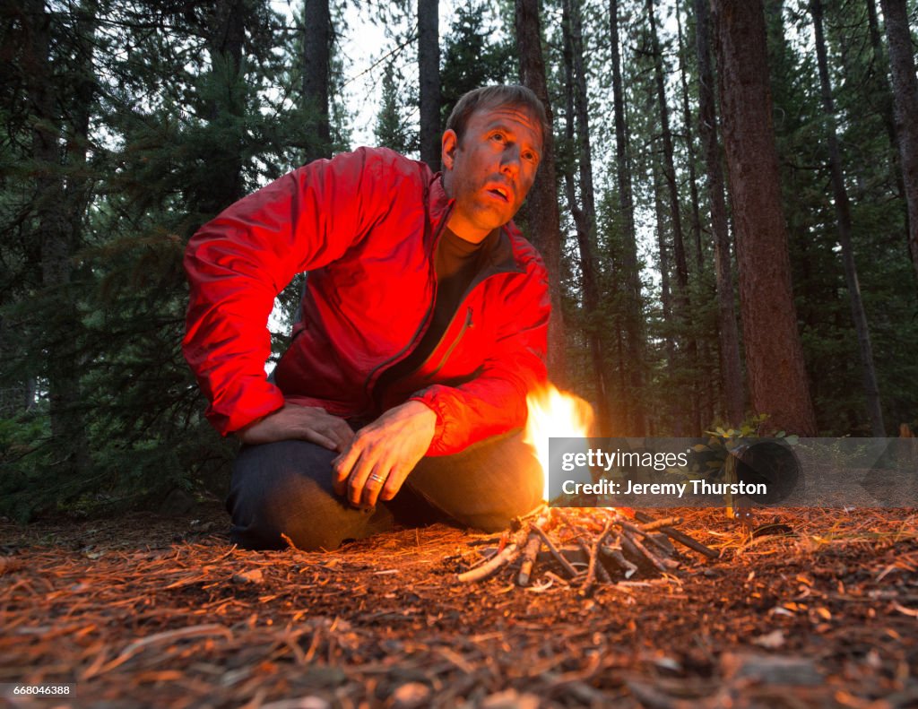 Man lost and alone in the woods sits by small fire as night approaches