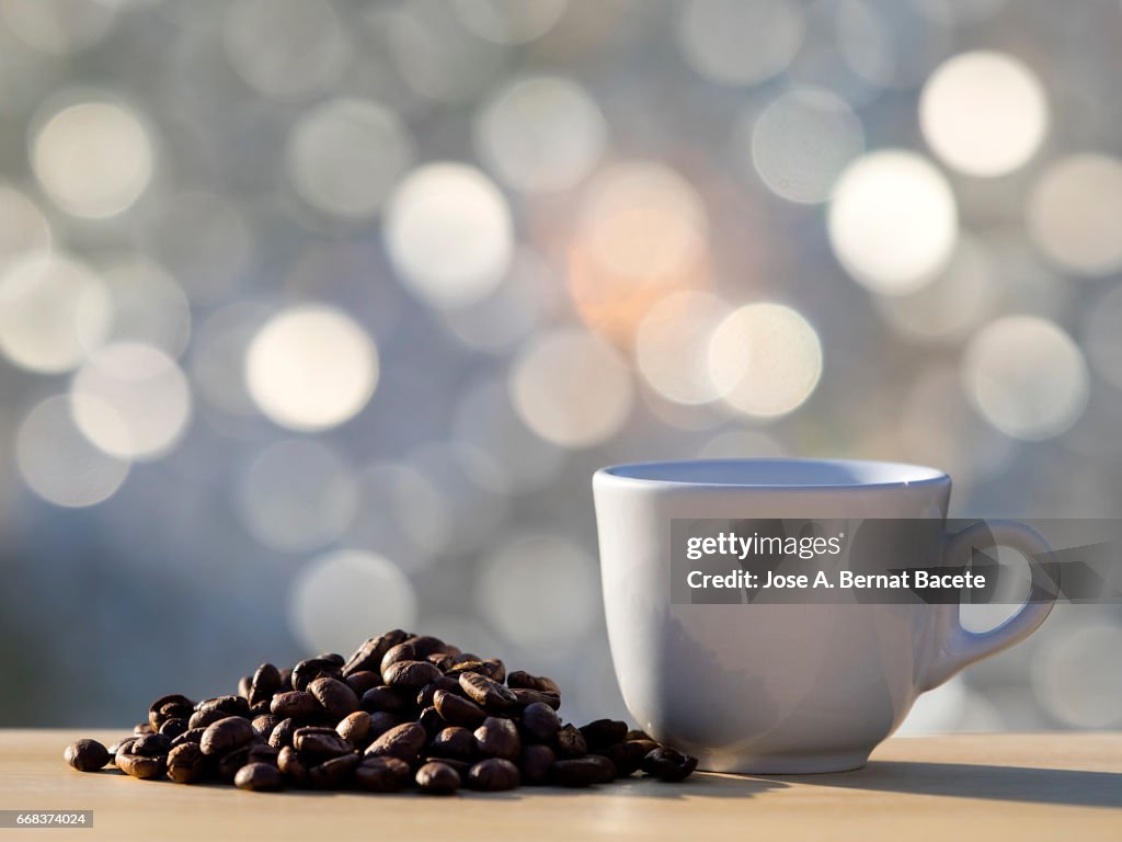 Heap of coffee beans toasted and a cup of coffee  on a table of wood illuminated by the light of the Sun