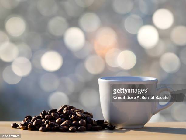 heap of coffee beans toasted and a cup of coffee  on a table of wood illuminated by the light of the sun - frescura stockfoto's en -beelden