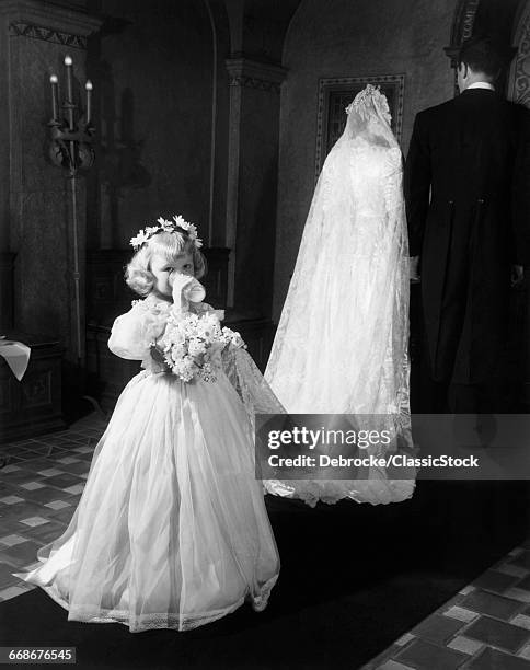 1950s LITTLE GIRL BRIDESMAID DRINKING GLASS OF MILK HOLDING BRIDAL VEIL TRAIN WALKING DOWN CHURCH AISLE LOOKING AT CAMERA