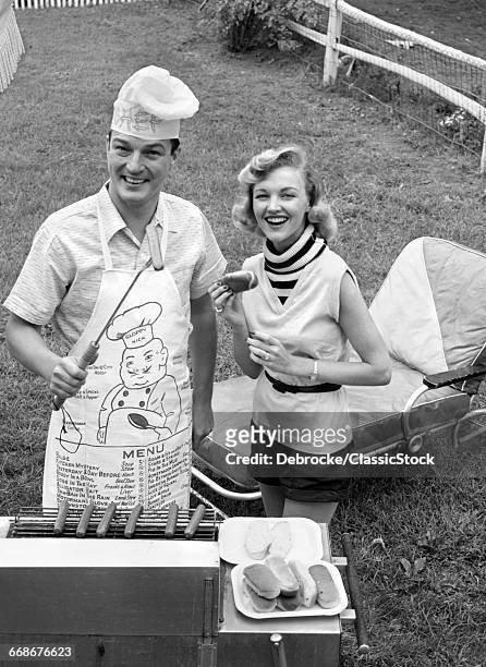 1950s 1960s COUPLE BACKYARD GRILLING LOOKING AT CAMERA COOKING HOT DOGS MAN WEARING APRON TOQUE & SKEWERED HOT DOG