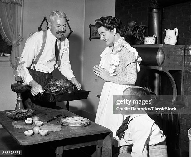 1890s 1900s MOCK TURN OF THE 20TH CENTURY THANKSGIVING DINNER FATHER TAKING TURKEY OUT OF STOVE SHOWING IT TO WIFE AND SON