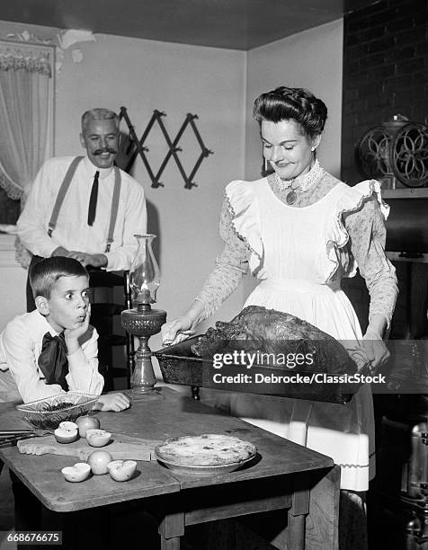 1950s MOCK TURN OF THE 20TH CENTURY THANKSGIVING DINNER SMILING MOTHER SHOWING ROAST TURKEY TO PLEASED HUSBAND AND SURPRISED SON