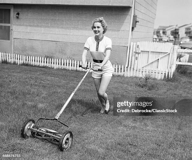 1950s SMILING BLOND WOMAN IN SHORTS MOWING LAWN WITH PUSH MOWER