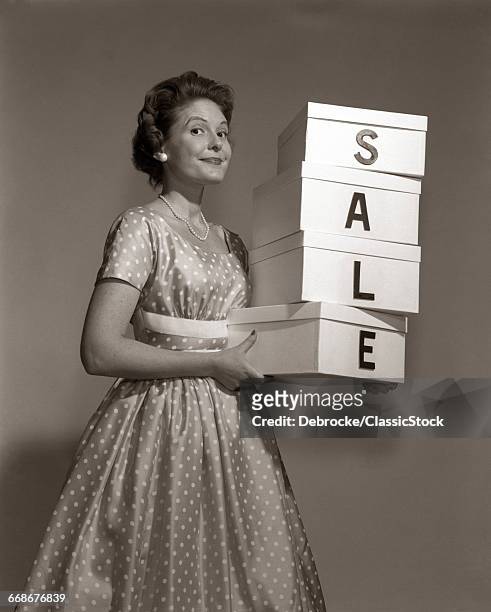 1960s WOMAN IN POLKA-DOT DRESS LOOKING AT CAMERA HOLDING A STACK OF 4 BOXES WITH ONE CHARACTER CENTERED ON EACH SPELLING SALE
