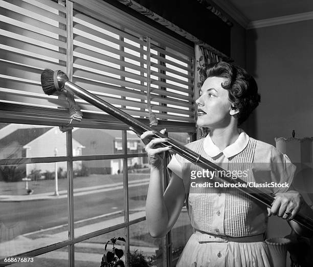 1950s WOMAN VACUUMING VENETIAN BLINDS ON WINDOW LOOKING OUT ONTO SUBURBAN STREET