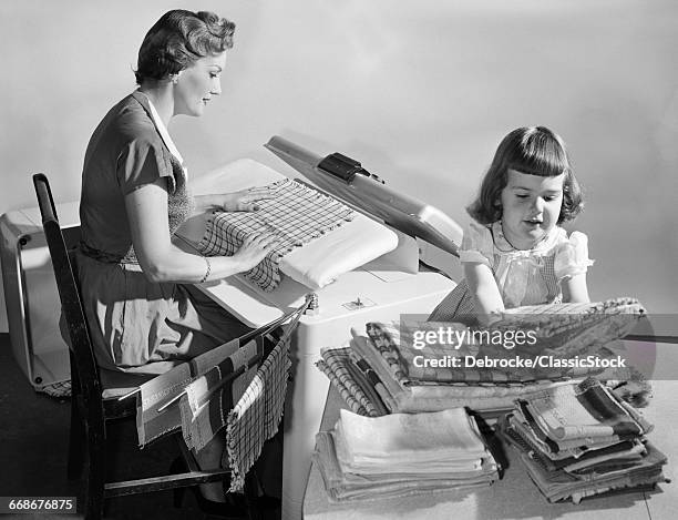 1950s HOUSEWIFE PRESSING LINENS ON MANGLE MACHINE AS DAUGHTER FOLDS & STACKS THEM ON TABLE