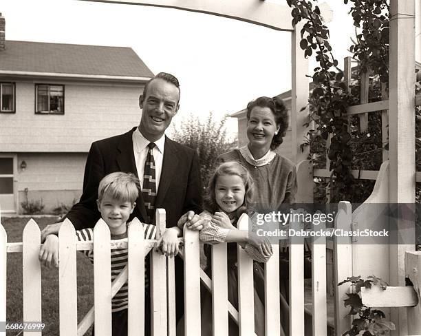 1950s SMILING FAMILY OF FOUR BEHIND PICKET FENCE IN BACKYARD LOOKING AT CAMERA