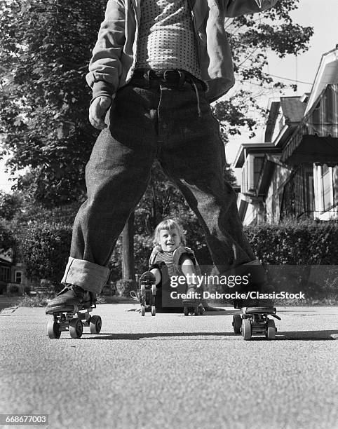 1950s LITTLE BOY AND GIRL PLAYING ON SIDEWALK WEARING METAL ROLLER SKATES AND BLUE JEANS GIRL HAS FALLEN ON HER BUTT