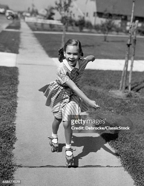 1950s LITTLE GIRL ROLLER-SKATING ON SIDEWALK TOWARDS AND LOOKING AT CAMERA