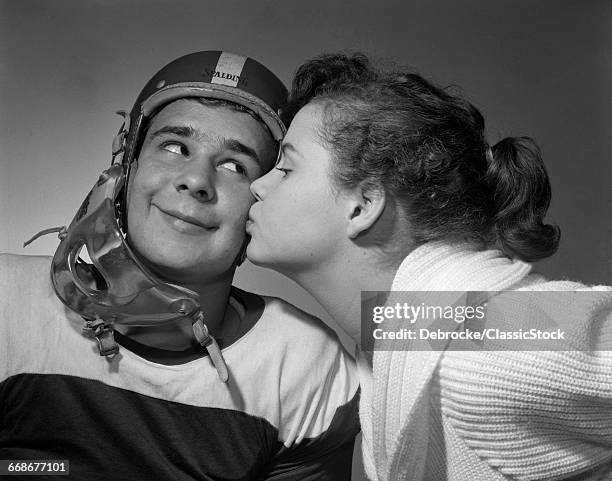 1950s TEEN GIRL WITH PONY TAIL KISSING BOY WEARING FOOTBALL HELMET ON THE CHEEK