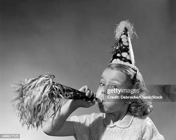 1950s GIRL WEARING PARTY HAT BLOWING INTO NOISE MAKER LOOKING AT CAMERA