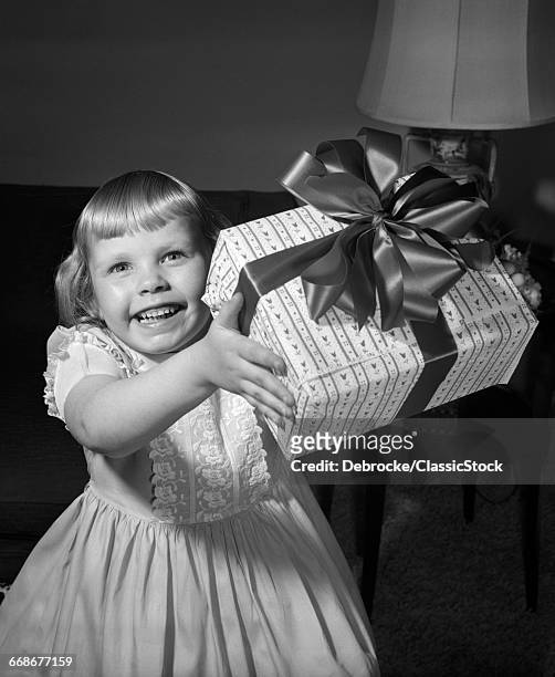 1950s SMILING GIRL HOLDING UP WRAPPED PRESENT EITHER GIVING OR RECEIVING A GIFT