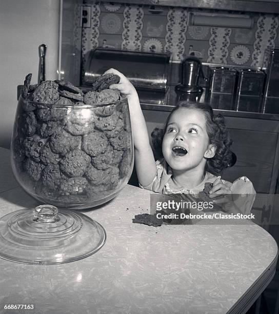 1950s WIDE-EYED GIRL WITH MOUTH AGAPE REACHING UP INTO COOKIE JAR TAKING ANOTHER CHOCOLATE CHIP COOKIE