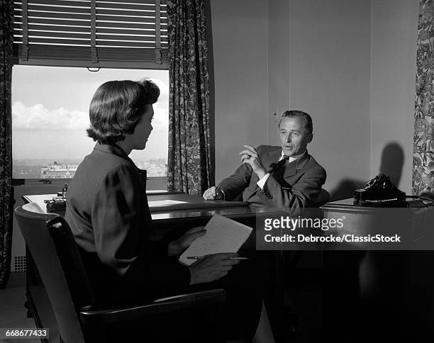 1950s BUSINESSMAN SITTING AT DESK SPEAKING DICTATING TO WOMAN STENOGRAPHER ALSO SEATED