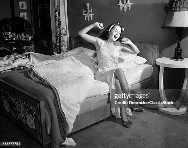 1950s SMILING YOUNG WOMAN IN NIGHTGOWN SLIPPERS SITTING EDGE BED LOOKING AT CAMERA YAWNING STRETCHING WAKING UP